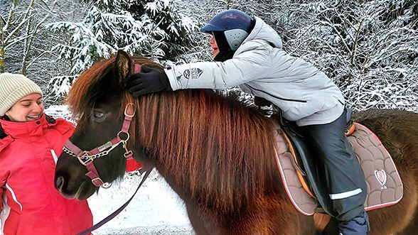 Ein Junge mit Down-Syndrom sitzt auf einem schwarzen Pferd. Er lehnt sich nach vorne und berührt die Ohren des Pferdes. Das Pferd wird von einer Person in einer roten Jacke geführt. 
