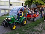 Ein Gartentraktor der Marke John Deere mit einem Anhänger. Im Anhänger sind acht Kinder drin. Fast alle lachen in die Kamera. Der Fahrer winkt fröhlich. Der Traktor ist dekoriert mit diversen Flaggen aus der Welt. 