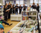 Ein Chor, bestehend aus 10 Personen, alle in schwarz gekleidet, singt in einer Bibliothek.