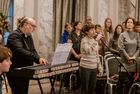 Eine Schülerin singt am Mikrofon. Daneben spielt eine Person am Keyboard. Im Hintergrund sehen wir den Chor.