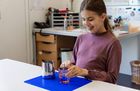 Eine Schülerin sitzt an einem Tisch. Vor ihr steht ein Glas mit einem Hilfsmittel, das hilft, die Flüssigkeit im Glas richtig zu dosieren.