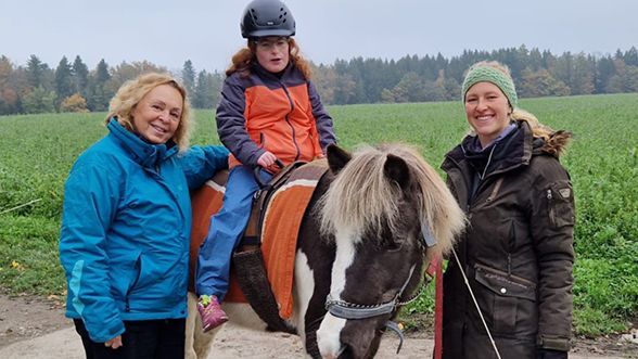 Ein Therapiepferd mit einem Kind auf dem Rücken. Das Pferd wird flankiert von zwei Frauen.